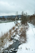 A small river in a regulated streambed and a wintry landscape with a church in the background. No people are walking along the hiking trail