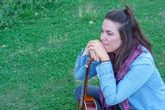 Attractive long-haired brunette girl dressed in blue sitting on the grass looking out of profile holding her ukulele