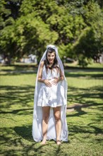 Pregnant bride in a white wedding dress holds her belly with her hands in a park. Happy