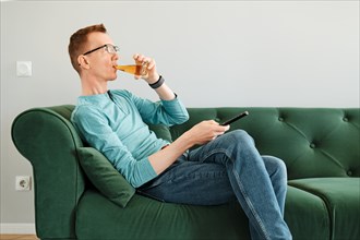 Man drinking juice and switching channels with remote control sitting on sofa in living room