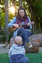 Woman playing ukulele smiling and a baby sitting on the grass watching her as a spectator