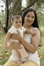 Portrait of attractive latin mother with her baby enjoying a sunny day in the park. Trees and greenery can be seen in the background