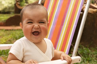 Close-up of baby girl laughing and looking up to camera outdoors in sunlight sitting on a colorful chair outdoors