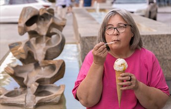 White-haired woman with glasses and pink T-shirt eating ice cream with a spoon sitting at a fountain