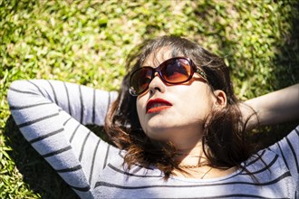 Overhead portrait of an Andean woman with sunglasses lying on the grass with lights and shadows filtered by the tree branches