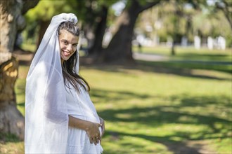 Pregnant bride in a white wedding dress holds her belly with her hands in a park. Happy expression