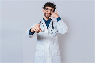 Smiling doctor making call gesture and pointing at the camera. Young doctor making call gesture isolated