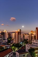 District of the city of Belo Horizonte in Minas Gerais with its buildings lit up by the sunset and with the full moon rising in the background