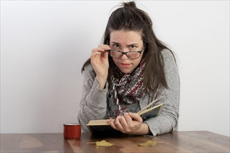 Young brunette woman with long hair and glasses reading a book holding her glasses and looking at the camera with a white background