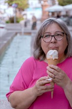 Close-up of a white-haired woman with glasses eating ice cream in front of a fountain
