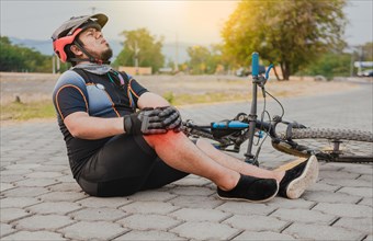 Male cyclist sitting on the pavement with knee pain. Concept of a cyclist with knee injury outdoors