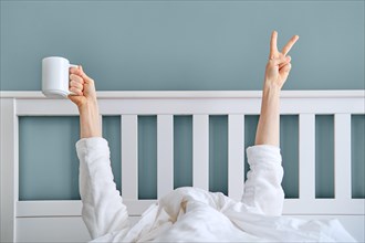 Man hiding under blanket showing hand with victory sign and holding cup of coffee. The early bird catches the worm concept