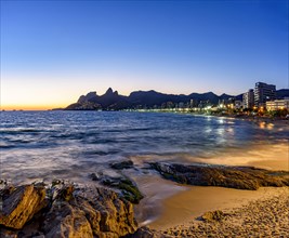 Dusk on Ipanema beach in Rio de Janeiro with the city lights on and the sky colored by the sunset reflecting in the sea water