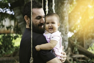 Portrait of father and baby smiling and looking at camera sitting in a hammock. Father's Day Concept. Sunset light