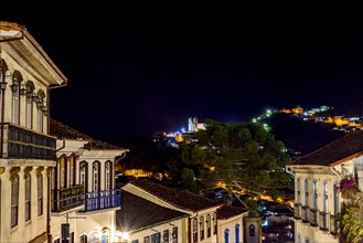 Facades of houses in colonial architecture on an old cobblestone street in the city of Ouro Preto illuminated at night with historic church in background