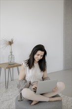 Young woman sitting comfortably with laptop on a stool in the living room