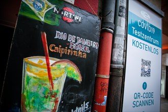 A signboard of a bar advertising Caipirinha cocktails stands next to the sign of a Covid-19 test centre in the old town of Duesseldorf