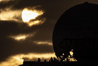 The silhouettes of people and the former listening station on Teufelsberg stand out against the setting sun in Berlin
