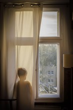 Symbolic photo on the subject of psychology in children. A boy stands at the window behind a curtain and looks out. Berlin