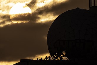 The silhouettes of people and the former listening station on Teufelsberg stand out against the setting sun in Berlin