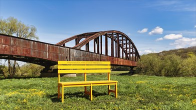 Oversized yellow wooden bench in front of old railway bridge