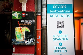 A board of a bar advertising Caipirinha cocktails stands next to the sign of a Covid-19 test centre in the old town of Duesseldorf