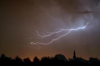 Forked lightning during thunderstorm at night over church tower and trees