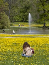 A young woman sits in a meadow with buttercups in front of a pond with fountain in the spa gardens of Bad Homburg vor der Hoehe