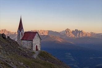 Little chapel and view over orange-lit Dolomite mountain range at sunset in summer