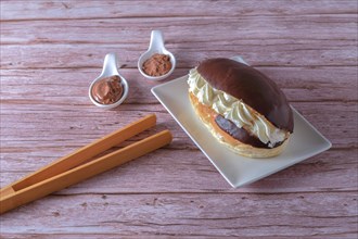 Delicious chocolate-covered cream eclair and white ceramic spoons with chocolate and cocoa on the side with wooden tongs