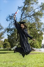 Happy graduated girl in a black gown jumping while holding her diploma
