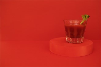 A glass of red cocktail with greens on the podium. Stylish alcohol tomato beverage Bloody Mary on a red background. Delicious drinks concept. Copy space