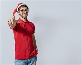 Christmas bearded man standing pointing at you isolated. Friendly guy in christmas hat with hands in his pocket pointing at you