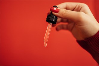 Woman with red manicure holding transparent serum or oil glass pipette. Bright red background. Mock up. Concept of cosmetic and selfcare. Copy space