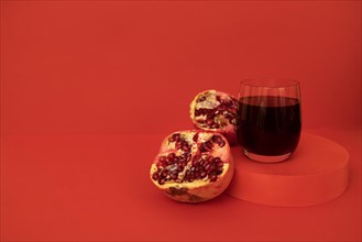 Glass of pomegranate juice on a podium with a pomegranate divided into pieces. Red bright background. Close up. Place for text. Healthy lifestyle concept