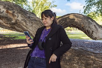 A woman is reading a text message on her mobile phone while leaning on a tree in the park