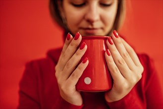 Young blondy girl in a red sweater holds a red cup in her hands. Close up of a beautiful bright red manicure. Valentine Day concept. Coffee break. Relax time