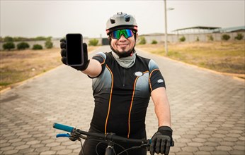 Cyclist in sportswear showing the phone screen. Male cyclist on the bike showing cell phone screen outdoors. Smiling cyclist showing an advertisement on cell phone