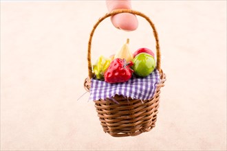 Hand holding a wicker basket with a plastic fruit on a white and wooden background
