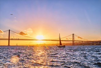 View of 25 de Abril Bridge famous tourist landmark of Lisbon connects Lisboa and Almada on Setubal Peninsula over Tagus river with tourist yacht silhouette at sunset and flying plane. Lisbon
