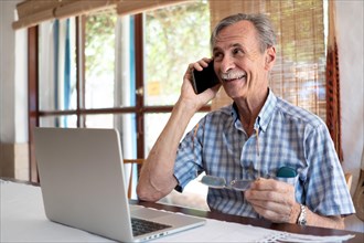 Mature business man smiling and talking on phone in front of laptop