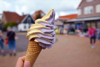 Close up of hand holding cone with mixed yellow and purple mango and berry soft serve ice cream with blurry city scene in background