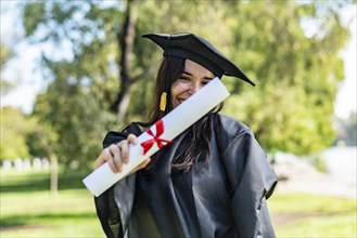 Happy caucasian graduated girl with long brown hair showing her diploma. She is wearing a bachelor gown and a black mortarboard