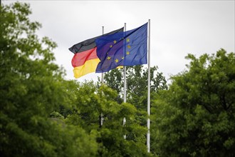 The flags of Europe and Germany fly in the wind behind trees. Berlin
