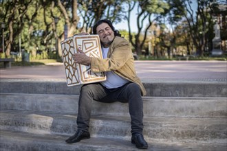 Hispanic musician sitting in a park hugging his flamenco box