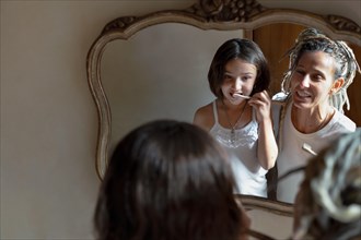 Girl looks in the mirror and brushes her teeth while her mother smiles at her. Image reflected in the bathroom mirror