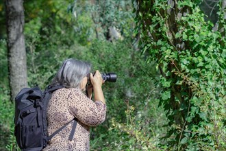 White-haired female photographer in profile and backpack view taking pictures of a ivy