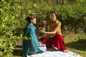 Concentrated female therapist holding a singing bowl performing therapy on a woman sitting in lotus position. Concept of sound healing therapy