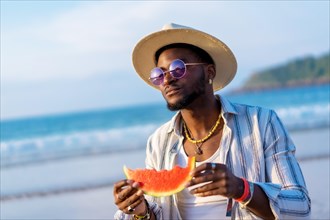 Portrait of a black ethnic man enjoy summer vacation at the beach eating a watermelon wearing a sunshade and sunglasses