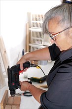 Woman see in profile drilling a wooden board with an electric drill on a white board in her workshop
